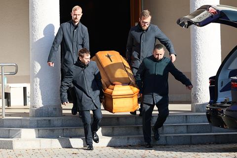 Four men in black coats carry a coffin to a waiting car in the sunshine.